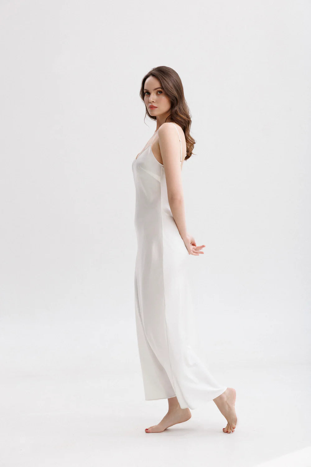 Woman wearing a long silk white slip dress on a spaghetti straps showing the side pose. 